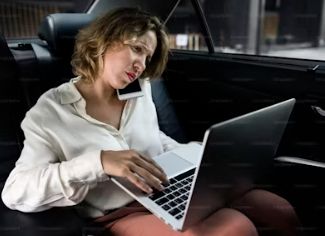A woman sitting in the back of a car, a phone held between cheek and shoulder, with a laptop on her knee. Creating goals can be really stressful! 